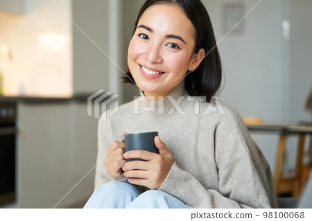 Portrait of smiling asian girl with cup of coffee, drinking hot tea and getting cozy at home. Warming up with beverage Portrait of smiling asian girl with cup of coffee, drinking hot tea and getting cozy at home. Warming up with beverage 98100068