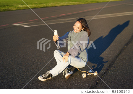 Beautiful korean girl takes selfie on smartphone, takes photo with her skateboard, enoys sunny day outdoors 98100143