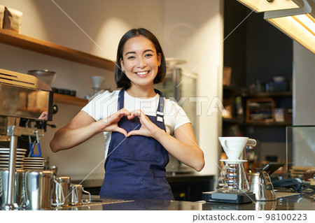 Smiling asian girl barista, shows heart sign, loves making coffee an serving clients, standing in uniform behind counter, work in cafe 98100223