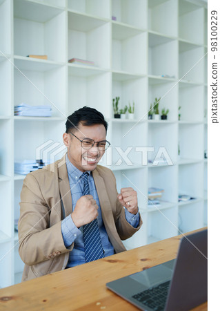 Portrait of an Asian male business owner standing with a computer showing happiness after a successful investment. Portrait of an Asian male business owner standing with a computer showing happiness after a successful investment. 98100229
