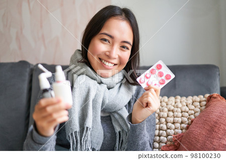 Portrait of happy smiling asian girl showing medication, sore throat spray and drugs from flu or cold, staying at home, wearing warm clothes Portrait of happy smiling asian girl showing medication, sore throat spray and drugs from flu or cold, staying at home, wearing warm clothes 98100230