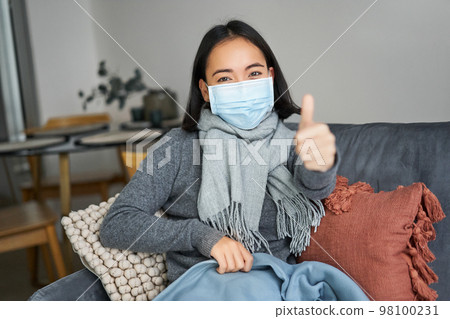 Covid-19 and health concept. Young asian woman in medical face mask, feels sick and unwell, catching flu, protecting others from influenza, sitting in living room covered with blanket Covid-19 and health concept. Young asian woman in medical face mask, feels sick and unwell, catching flu, protecting others from influenza, sitting in living room covered with blanket 98100231