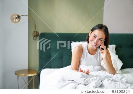 Smiling korean girl talks on mobile phone and lying in bed. Cute woman answers telephone call, holds smartphone, relaxing in her bed 98100294