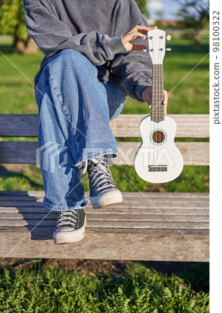 Cropped shot of young girl in sneakers and jeans, hands holding ukulele musical instrument while she sits on bench in green sunny park Cropped shot of young girl in sneakers and jeans, hands holding ukulele musical instrument while she sits on bench in green sunny park 98100322