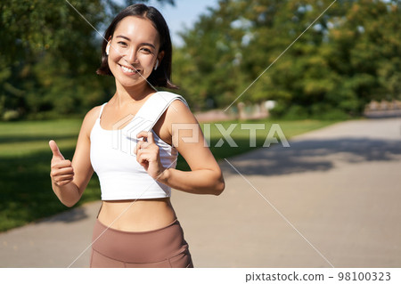 Smiling fitness girl shows thumb up, stands with towel after good workout in park, jogging and looking happy Smiling fitness girl shows thumb up, stands with towel after good workout in park, jogging and looking happy 98100323