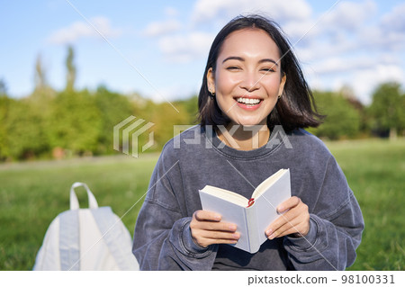 Beautiful asian girl sitting in park on grass, reading and smiling. Woman with book enjoying sunny day outdoors Beautiful asian girl sitting in park on grass, reading and smiling. Woman with book enjoying sunny day outdoors 98100331