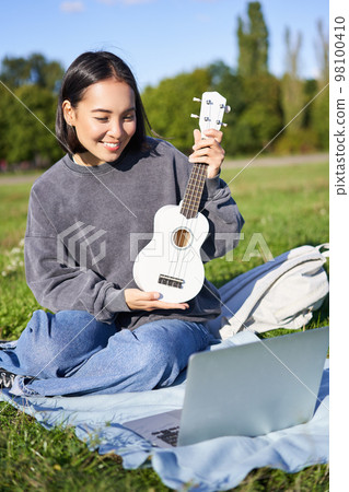 Vertical shot of happy asian girl plays instrument, shows her ukulele at laptop camera, video chats about music, teaches how to play, sits in park outdoors Vertical shot of happy asian girl plays instrument, shows her ukulele at laptop camera, video chats about music, teaches how to play, sits in park outdoors 98100410