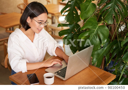Working female entrepreneur sitting in cafe with laptop, typing on computer, drinking coffee Working female entrepreneur sitting in cafe with laptop, typing on computer, drinking coffee 98100450