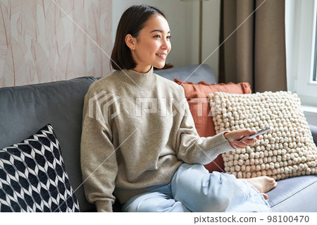 Beautiful asian girl in cozy home, watching television, holding remote and switching channels, sitting on sofa Beautiful asian girl in cozy home, watching television, holding remote and switching channels, sitting on sofa 98100470