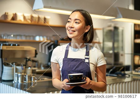 Portrait of smiling asian woman in apron, barista giving you cup of coffee, working in cafe, serving drinks 98100617