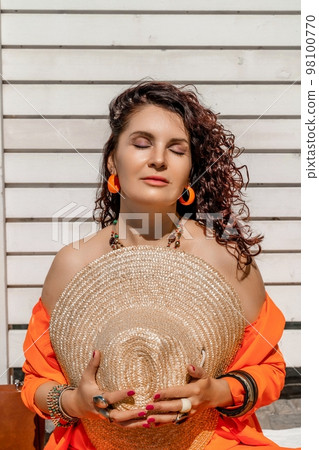 Stylish woman in an orange suit holds a straw hat in her hands. On the hands are jewelry rings and bracelets, on a white striped background. 98100770