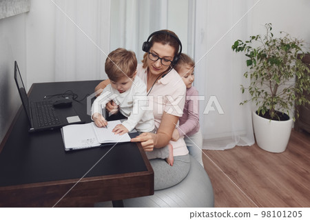 Mother sits on a fitness ball in headphones and works at home with her children during quarantine Mother sits on a fitness ball in headphones and works at home with her children during quarantine 98101205