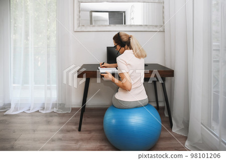 Woman sits on a fitness ball in headphones and works at home during quarantine 98101206
