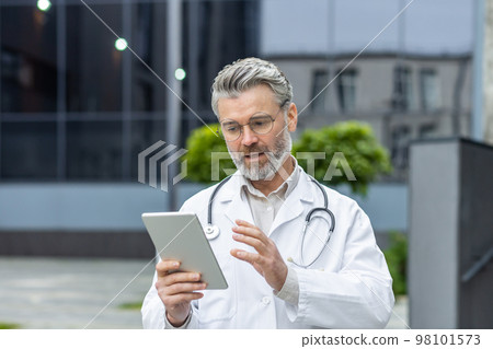 Serious mature doctor in medical gown outside modern clinic using tablet computer for video call, man with stethoscope consults patient online remotely. Serious mature doctor in medical gown outside modern clinic using tablet computer for video call, man with stethoscope consults patient online remotely. 98101573