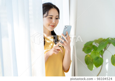 A young woman operating a smartphone by the window A young woman operating a smartphone by the window 98104524