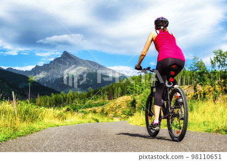 Woman riding mountain bike on road in forest with hill at background in High Tatras mountains, Slovakia 98110651
