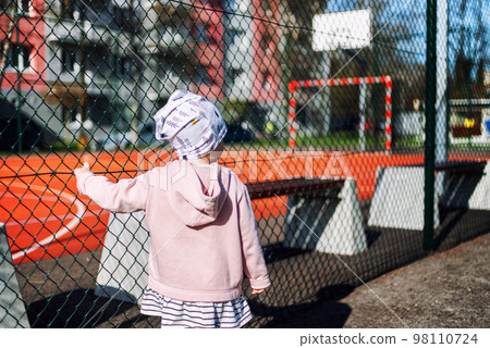 Small girl toddler holding fence and watching on closed empty playground Small girl toddler holding fence and watching on closed empty playground 98110724