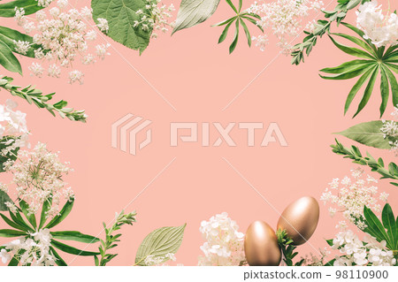 Golden Easter eggs in assorted floral and jungle tree green leaves and white flowers frame border on peach pink. Natural sustainable environment friendly cosmetics mockup flat lay with copy space 98110900