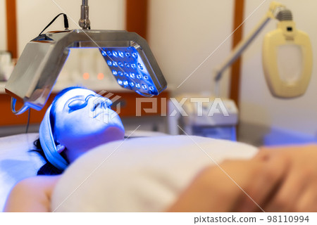 Facial treatment with led therapy. Girl on a light therapy procedure. LED lamp with blue light. Woman in protective glasses. Facial treatment with led therapy. Girl on a light therapy procedure. LED lamp with blue light. Woman in protective glasses. 98110994