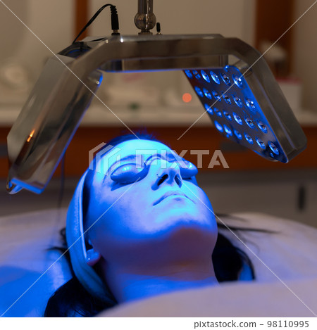 Young woman having blue LED light facial therapy treatment in beauty salon. Young woman having blue LED light facial therapy treatment in beauty salon. 98110995