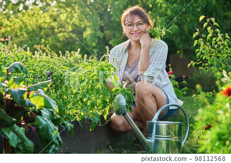 Middle-aged woman with watering can near garden bed with spicy fragrant herbs Middle-aged woman with watering can near garden bed with spicy fragrant herbs 98112568