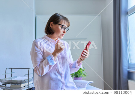 Middle aged woman sitting at workplace using smartphone in home office Middle aged woman sitting at workplace using smartphone in home office 98113004