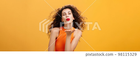 Flirty confident and sensual stylish woman with curly hairstyle and red lipstick folding lips holding palm near mouth sending kiss while hair flicks on air being in romantic mood over orange wall 98115329