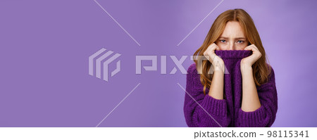 Gloomy upset redhead girlfriend pulling collar of purple sweater on nose, frowning and squinting offended, being aggrieved and resent, sulking moody and displeased over violet background 98115341