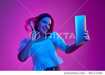 Portrait of young emotive girl posing in white blouse, haing online video call on tablet over pink background in neon light. Concept of emotions, youth, lifestyle Portrait of young emotive girl posing in white blouse, haing online video call on tablet over pink background in neon light. Concept of emotions, youth, lifestyle 98115360