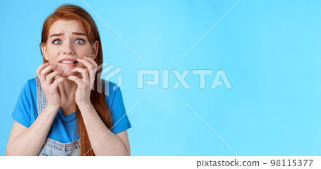 Close-up intense panicking redhead woman watching scary movie feel pressured, biting lip worried, hold hands near mouth self-soothing trying calm down, stare camera anxious, blue background 98115377
