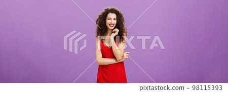 Gorgeous lady in red feeling shy and awkward being invited for dance standing timid and feminine over purple background touching strand of curly hair smiling broadly and crossing arm over chest Gorgeous lady in red feeling shy and awkward being invited for dance standing timid and feminine over purple background touching strand of curly hair smiling broadly and crossing arm over chest 98115393