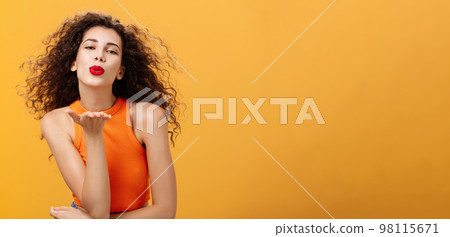 Flirty confident and sensual stylish woman with curly hairstyle and red lipstick folding lips holding palm near mouth sending kiss while hair flicks on air being in romantic mood over orange wall 98115671
