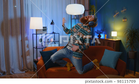 Man in headphones dancing on sofa at home listening favorite music, playing on imaginary guitar 98115709