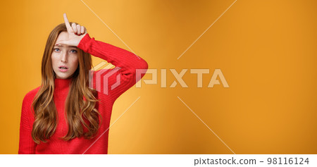 Arrogant and self-satisfied redhead woman humiliating person showing loser sign on forehead mocking and disdain rival standing confident and snobbish over orange background 98116124