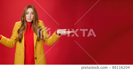 Who new it happen. Innocent and cute redhead unaware girl with freckles in yellow coat shrugging with hands spread sideways, pouting sad and gloomy, being uanware and clueless over red wall 98116204