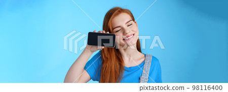 Carefree tender redhead european female tilt head, smiling closed eyes, hold smartphone horizontally, show loving girlfriend photos, romantic video online, stand blue background cheerful Carefree tender redhead european female tilt head, smiling closed eyes, hold smartphone horizontally, show loving girlfriend photos, romantic video online, stand blue background cheerful 98116400