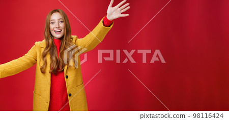 Hey come and hug me friend. Charming playful and emotional redhead female extanding palms forward to cuddle and greet, giving warm welcome smiling broadly posing in yellow coat over red wall 98116424