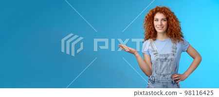 Let me show you cool product. Attractive cheerful confident redhead female model curly hairstyle present customers object hold palm blank blue copy space advertising standing blue background 98116425