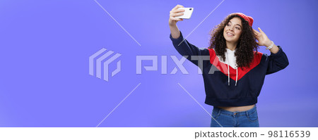 Cute girl making selfie to memorize trip standing in warm winter beanie and sweatshirt posing as pulling hand with smartphone and smiling at device screen to take pretty shot and post internet Cute girl making selfie to memorize trip standing in warm winter beanie and sweatshirt posing as pulling hand with smartphone and smiling at device screen to take pretty shot and post internet 98116539