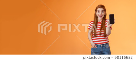 Excited cute outgoing redhead female impressed showing app, hold smartphone introduce gadget feature, smiling surprised, look enthusiastic like application, brag game score, orange background 98116682
