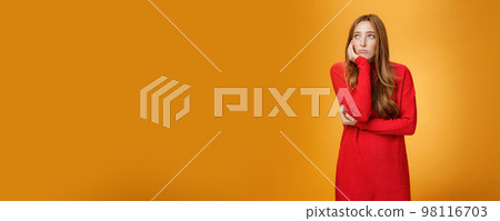 Sorrow and upset lonely woman leaning head on palm in upset pose looking gloomy at upper left corner as remembering unhappy story or being bored standing uneasy over orange wall 98116703