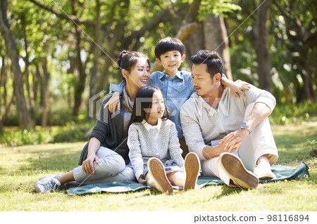 asian family with two children having fun sitting on grass talking chatting outdoors in park 98116894