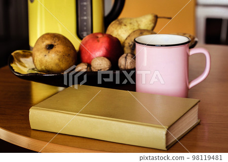 Autumnal still life: pink cup, yellow teapot, bowl of fruit, hardcover book lying on wooden table. Retro and modern housewares, utensils. Reading at cozy sweet home atmosphere. Lifestyles concept. Autumnal still life: pink cup, yellow teapot, bowl of fruit, hardcover book lying on wooden table. Retro and modern housewares, utensils. Reading at cozy sweet home atmosphere. Lifestyles concept. 98119481