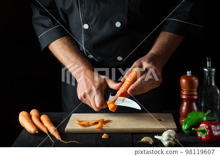 Professional chef peels carrots for vegetable soup in a restaurant kitchen. Close-up - hands of the cook during work. Carrot diet Professional chef peels carrots for vegetable soup in a restaurant kitchen. Close-up - hands of the cook during work. Carrot diet 98119607