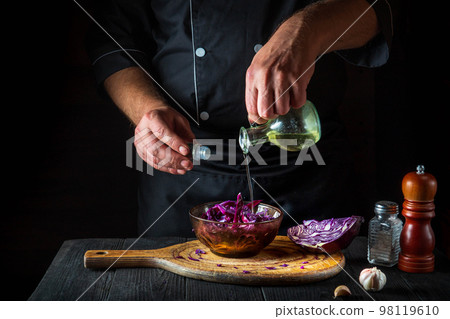 A professional chef pours olive oil into a fresh red cabbage salad. Cooking healthy food in restaurant kitchen on vintage table. 98119610