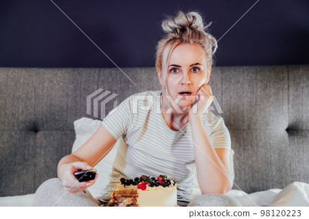 Emotionally stressed, shocked woman eating cake and holding a remote control while watching TV in bed. Scary horror movie or terrible news, scare in her eyes Overeating, Jamming of negative emotions. Emotionally stressed, shocked woman eating cake and holding a remote control while watching TV in bed. Scary horror movie or terrible news, scare in her eyes Overeating, Jamming of negative emotions. 98120223