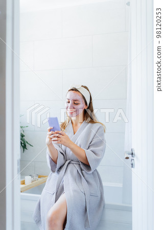 Smiling woman with facial pink clay mask on the face using smartphone at home bath enjoying relaxation and spa beauty treatment. Beauty blogger creating content. Vertical. Selective focus, copy space Smiling woman with facial pink clay mask on the face using smartphone at home bath enjoying relaxation and spa beauty treatment. Beauty blogger creating content. Vertical. Selective focus, copy space 98120253