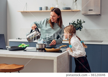 Woman cooks a meal with her curious schoolgirl daughter nearby 98120846