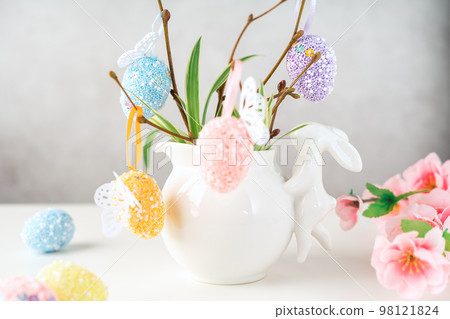 Home interior with easter decor. Vase with willow tree branches with Easter eggs and bunny on white table and background with copy space Home interior with easter decor. Vase with willow tree branches with Easter eggs and bunny on white table and background with copy space 98121824