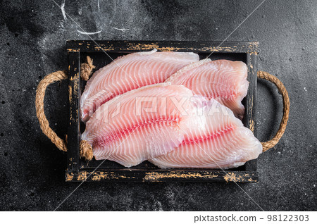 Fresh tilapia white fish fillet in a wooden tray. Black background. Top view Fresh tilapia white fish fillet in a wooden tray. Black background. Top view 98122303